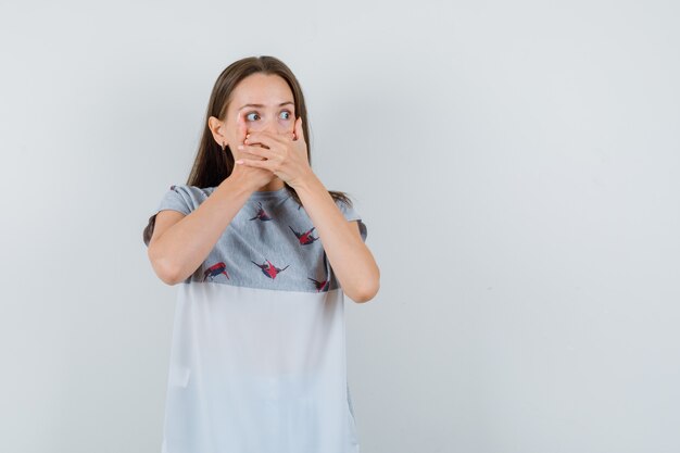 Niña cubriendo la boca con fuerza con las manos en camiseta y mirando asustado, vista frontal.