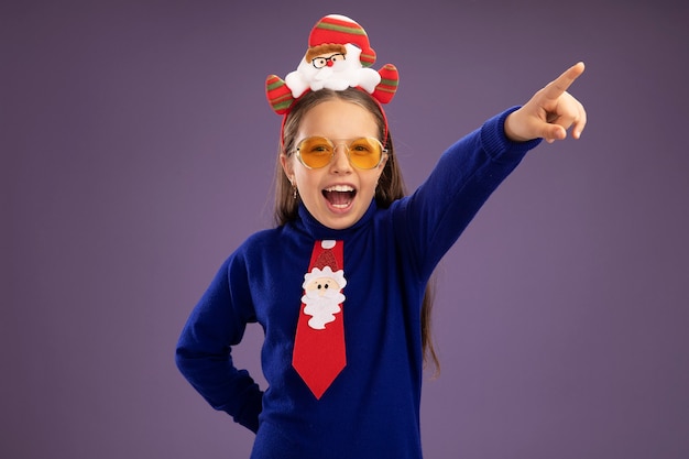 Niña de cuello alto azul con corbata roja y borde de navidad divertido en la cabeza feliz y emocionado mirando a la cámara apuntando con el dedo índice hacia el lado de pie sobre fondo púrpura