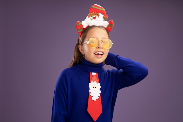 Niña de cuello alto azul con corbata roja y borde de navidad divertido en la cabeza mirando a cámara con cara feliz sonriendo alegremente de pie sobre fondo púrpura