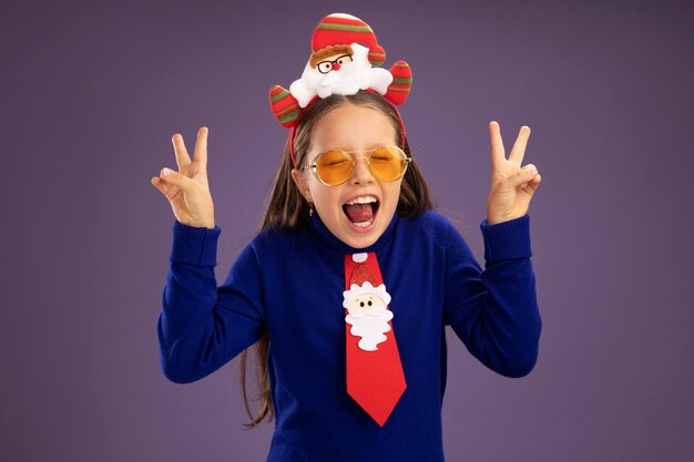 Foto gratuita niña de cuello alto azul con corbata roja y divertido borde navideño en la cabeza, feliz y emocionado mostrando el signo v de pie sobre fondo púrpura