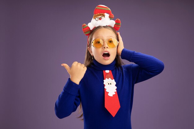 Foto gratuita niña de cuello alto azul con corbata roja y divertido borde navideño en la cabeza mirando a la cámara asombrado apuntando con el pulgar hacia el lado de pie sobre fondo púrpura