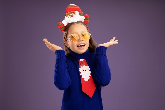 Niña de cuello alto azul con corbata roja y divertido borde navideño en la cabeza mirando a la cámara feliz y emocionado con los brazos levantados sobre fondo púrpura