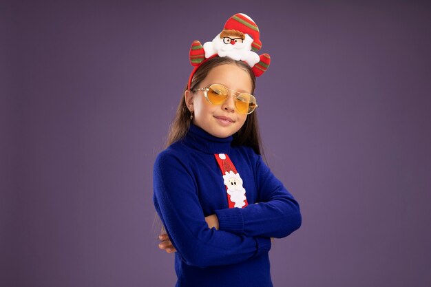 Niña de cuello alto azul con corbata roja y divertido borde navideño en la cabeza mirando a la cámara sonriendo confiado con los brazos cruzados sobre fondo púrpura