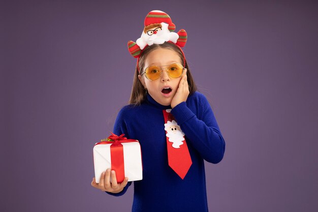 Niña de cuello alto azul con corbata roja y divertido borde navideño en la cabeza sosteniendo un presente mirando a la cámara asombrado de pie sobre fondo púrpura
