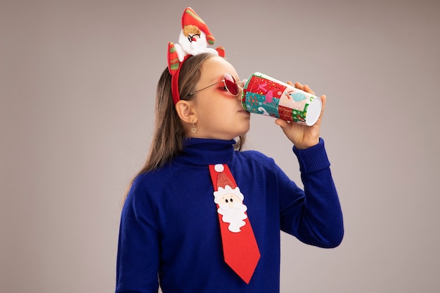 Niña de cuello alto azul vistiendo gracioso borde navideño en la cabeza bebiendo de coloridos vasos de papel de pie sobre la pared blanca