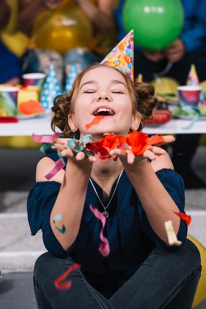 Foto gratuita una niña de cumpleaños con sombrero de fiesta en la cabeza con confeti cayendo en la mano