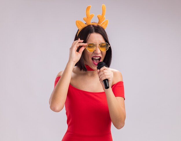 Niña con diadema de cuernos de reno y gafas sosteniendo micrófono agarrando gafas cantando con los ojos cerrados aislado sobre fondo blanco con espacio de copia