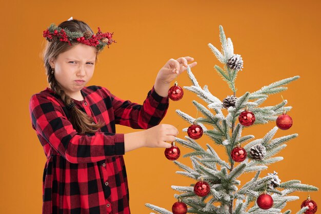 Niña disgustada vistiendo corona de navidad en vestido marcado decorando el árbol de navidad con el ceño fruncido de pie sobre la pared naranja