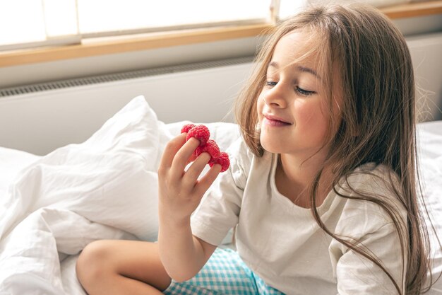 Niña divertida con frambuesas en los dedos en la cama por la mañana