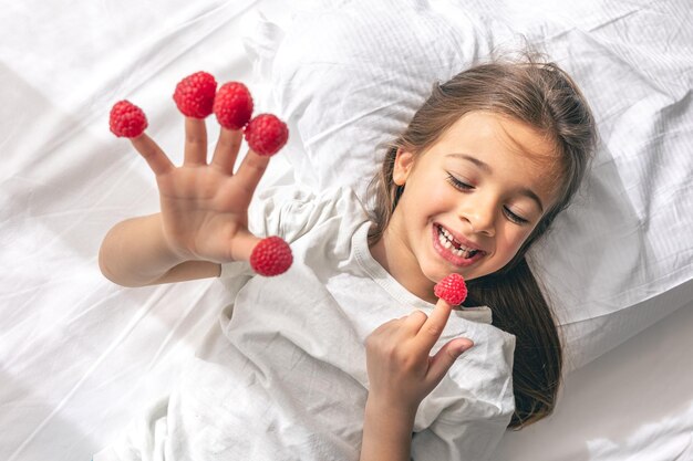 Niña divertida con frambuesas en los dedos en la cama por la mañana