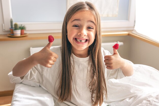 Niña divertida con frambuesas en los dedos en la cama por la mañana