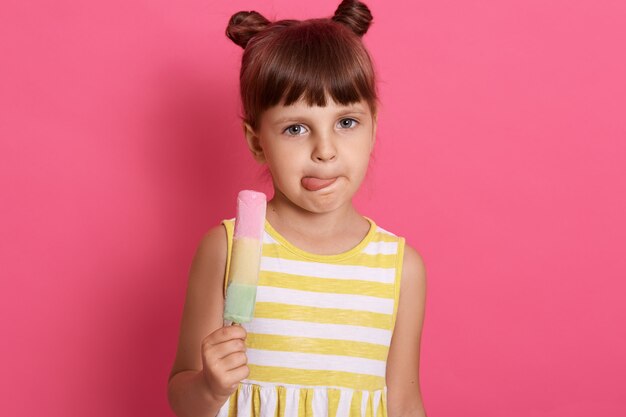 Niña divertida mientras se lame los labios, encantadora niña disfrutando de comer sorbete de frutas, posando aislada sobre pared rosada.