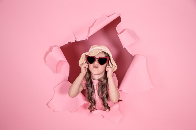 Niña divertida que se asoma por un agujero en un sombrero de playa y gafas en forma de corazón sobre un fondo de color, lugar para texto, fotografía de estudio