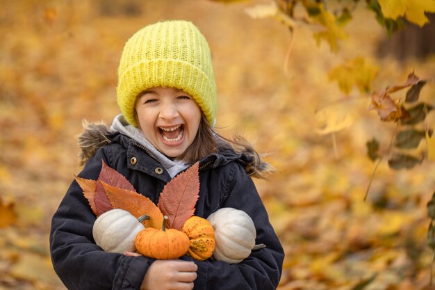 Niña divertida con un sombrero amarillo con pequeñas calabazas en el bosque de otoño sobre un fondo borroso, copie el espacio.