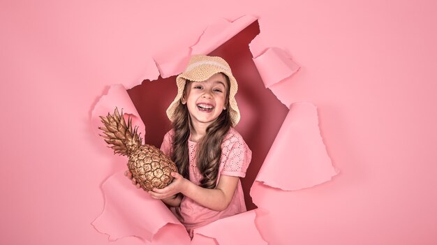 Niña divertida con un sombrero de playa y con color dorado piña, sobre un fondo de color rosa, asomando desde el agujero en el fondo, fotografía de estudio, espacio para texto