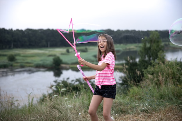 Una niña divertida sopla pompas de jabón en el verano en un campo, actividades de verano al aire libre.