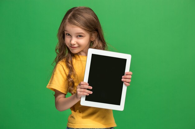 Niña divertida con tableta sobre fondo verde de estudio. Ella muestra algo y apunta a la pantalla.