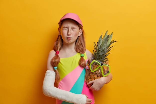 Niña con dos colas de caballo, cara pecosa, cierra los ojos y hace una mueca divertida, se divierte durante las vacaciones de verano, usa traje de baño y gorra, sostiene piña con máscara de snorkel, brazo roto en yeso