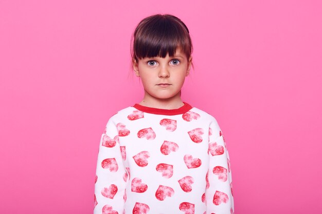 Niña en edad preescolar asustada mirando a la cámara con grandes ojos llenos de miedo, vestida con atuendo casual, niña de cabello oscuro con expresión de shock, aislada sobre una pared rosa.