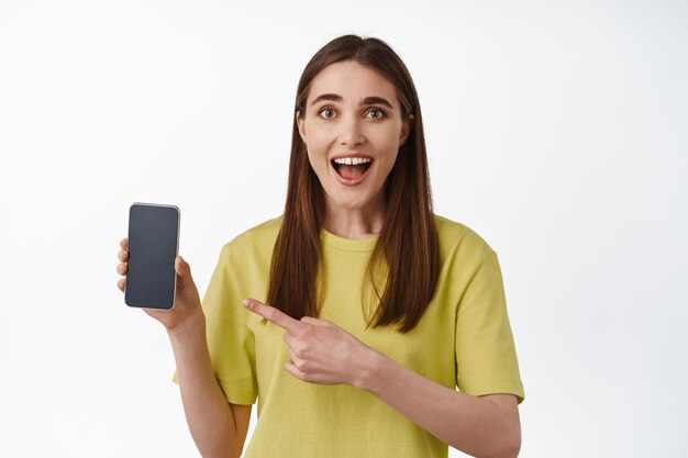 Niña emocionada revisando la aplicación, apuntando a la pantalla del teléfono inteligente, mostrando la pantalla o la interfaz de la aplicación en el teléfono móvil, de pie contra un fondo blanco.