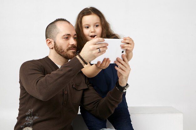 Niña emocional abriendo la boca ampliamente, asombrada con la nueva tableta digital, usándola con su moderno padre joven con barba