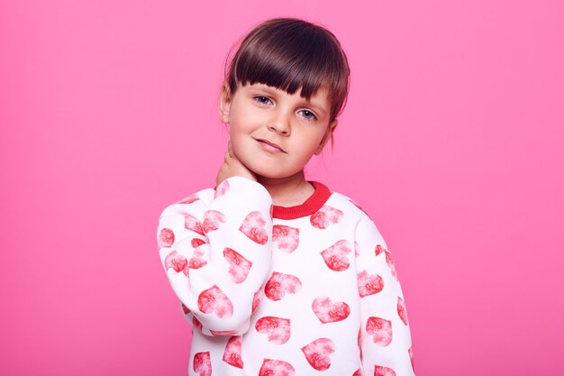 Niña encantadora con cabello oscuro que sufre de dolor, mirando a la cámara con el ceño fruncido, manteniendo las manos en el cuello, aislado sobre una pared rosa.