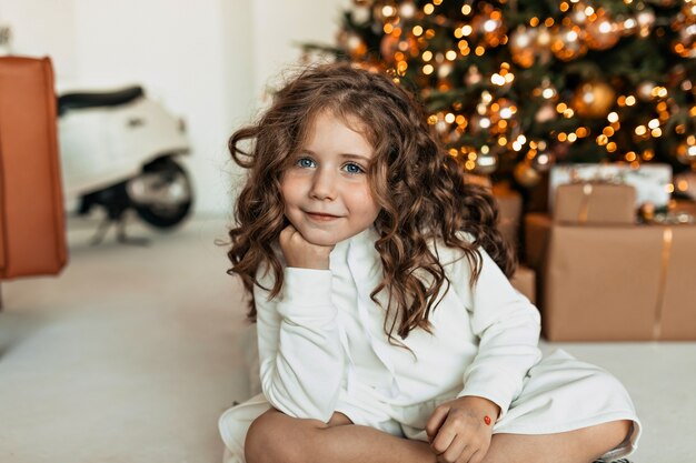 Niña encantadora de ensueño con rizos vistiendo un suéter de punto blanco sentado delante del árbol de Navidad y esperando a Santa