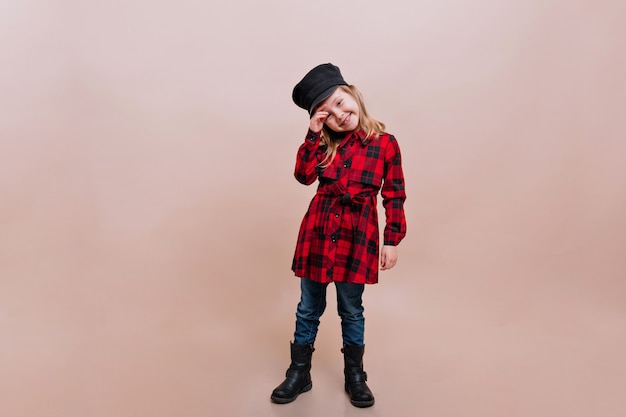 Niña encantadora vestida con camisa a cuadros, jeans y gorra elegante posa en una pared aislada con verdaderas emociones felices