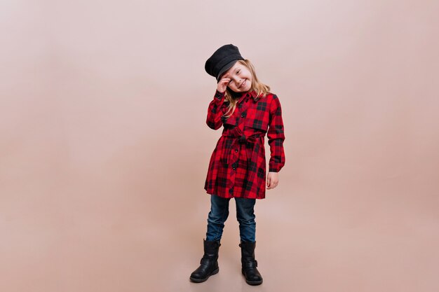 Niña encantadora vestida con camisa a cuadros, jeans y gorra elegante posa en una pared aislada con verdaderas emociones felices