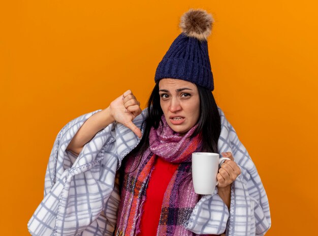 Niña enferma caucásica joven disgustada con gorro de invierno y bufanda envuelta en cuadros sosteniendo una taza de té mostrando el pulgar hacia abajo aislado en la pared naranja