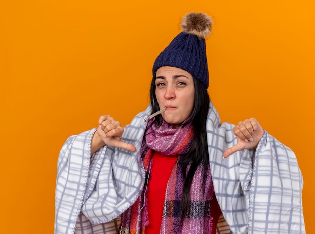 Niña enferma caucásica joven disgustada con gorro de invierno y bufanda envuelta en cuadros sosteniendo el termómetro en la boca mirando a la cámara mostrando los pulgares hacia abajo aislado sobre fondo naranja