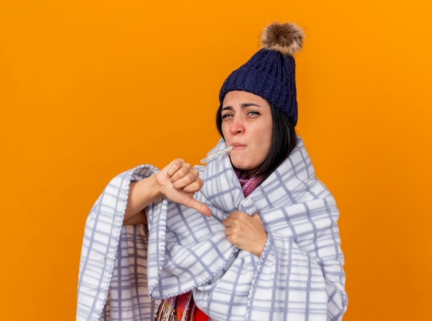 Foto gratuita niña enferma caucásica joven disgustada con gorro de invierno y bufanda envuelta en plaid sosteniendo el termómetro en la boca agarrando plaid mirando a cámara aislada sobre fondo naranja