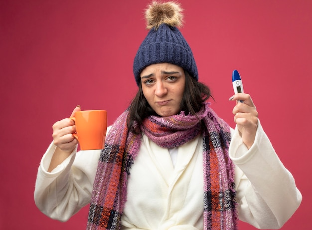 Foto gratuita niña enferma caucásica joven disgustada con sombrero de invierno bata y bufanda sosteniendo una taza de té y un termómetro mirando a la cámara aislada sobre fondo carmesí