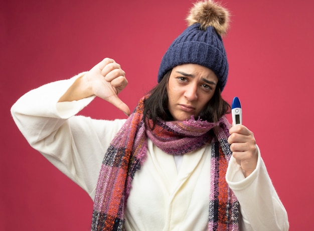 Foto gratuita niña enferma caucásica joven disgustada vistiendo bata gorro de invierno y bufanda sosteniendo el termómetro mirando a la cámara mostrando el pulgar hacia abajo aislado sobre fondo carmesí