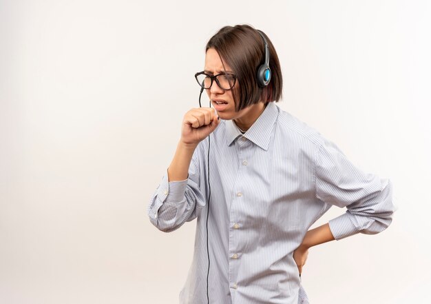 Niña enferma del centro de llamadas con gafas y auriculares manteniendo el puño cerca de la boca tosiendo y poniendo la mano en la cintura aislada en blanco con espacio de copia