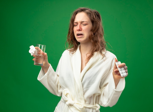 Niña enferma estornuda vistiendo túnica blanca sosteniendo un vaso de agua con pastillas aisladas en verde