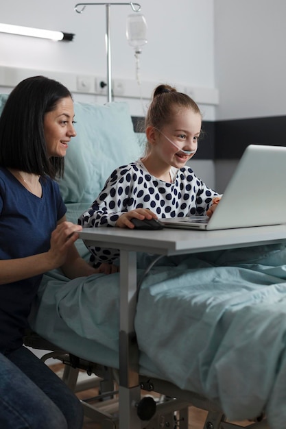 Niña enferma feliz jugando juegos en la computadora portátil mientras la madre se sienta a su lado dentro de la sala de pediatría del hospital. Niño alegre bajo tratamiento jugando en la cama del paciente mientras los padres la cuidan.