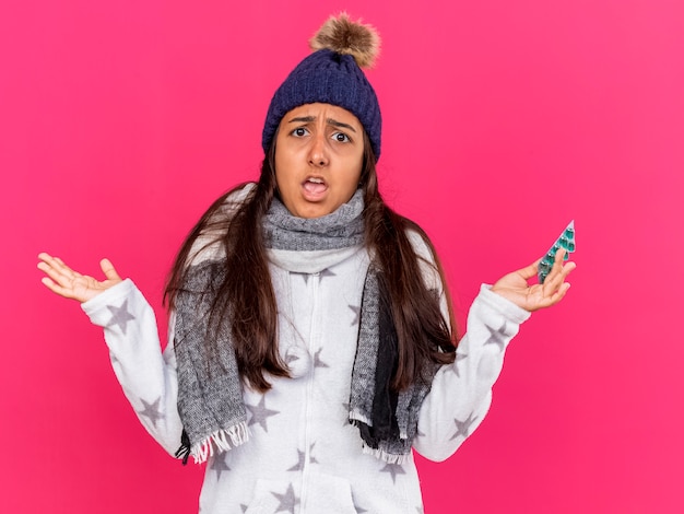 Niña enferma joven disgustada con sombrero de invierno con bufanda sosteniendo pastillas y extendiendo las manos aisladas sobre fondo rosa