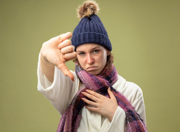 Niña enferma joven insatisfecha con bata blanca y gorro de invierno con bufanda mostrando el pulgar hacia abajo aislado en verde oliva