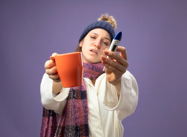 Niña enferma joven insatisfecha con bata blanca y gorro de invierno con bufanda sosteniendo una taza de té con termómetro aislado en púrpura