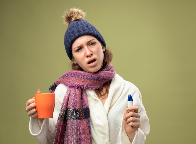 Niña enferma joven insatisfecha con bata blanca y gorro de invierno con bufanda sosteniendo una taza de té con termómetro aislado en verde oliva con espacio de copia