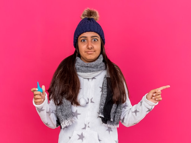 Foto gratuita niña enferma joven sorprendida con sombrero de invierno con bufanda sosteniendo pastillas y puntos en el lado aislado en rosa