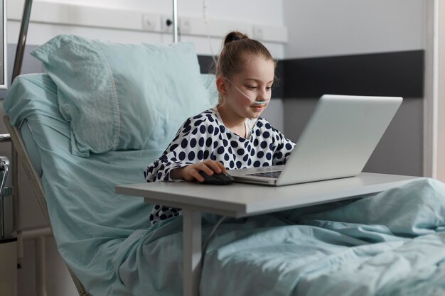 Niña enferma jugando juegos en la computadora portátil mientras está hospitalizada en la sala de pediatría de la clínica de salud. Niño enfermo descansando en la cama del paciente mientras disfruta de los juegos en la computadora moderna.