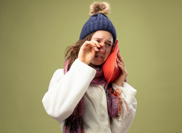 Niña enferma llorando con bata blanca y gorro de invierno con bufanda poniendo una bolsa de agua caliente en la mejilla limpiando los ojos con la mano aislada en verde oliva