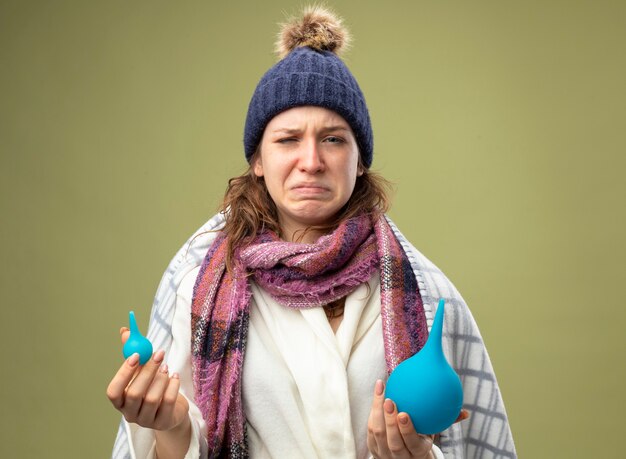 Niña enferma llorando vistiendo túnica blanca y gorro de invierno con bufanda envuelta en cuadros con enemas aislados en verde oliva