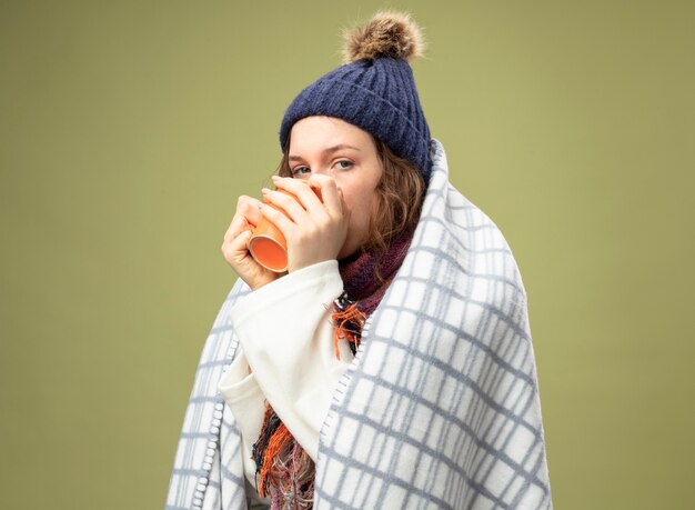 Niña enferma mirando al frente vistiendo túnica blanca y gorro de invierno con bufanda envuelta en plaid bebe té aislado en verde oliva