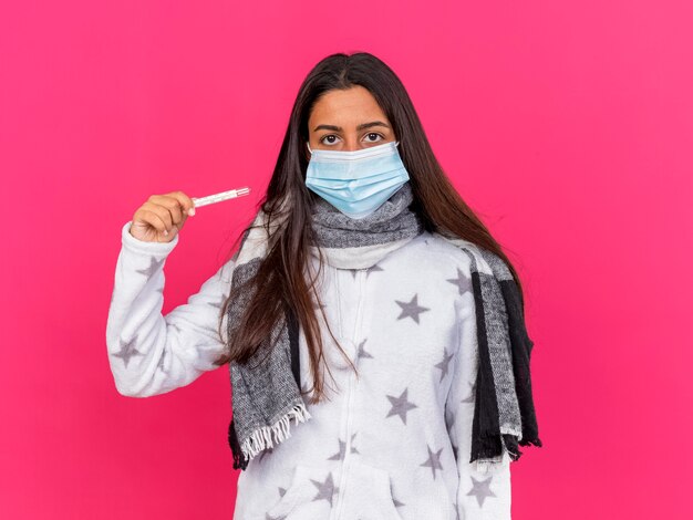 Niña enferma mirando a la cámara con máscara médica con bufanda sosteniendo termómetro aislado sobre fondo de color rosa