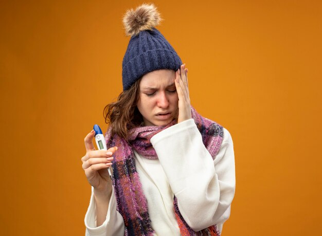 Niña enferma molesta con los ojos cerrados con bata blanca y gorro de invierno con bufanda sosteniendo el termómetro poniendo la mano en la mejilla aislada en naranja