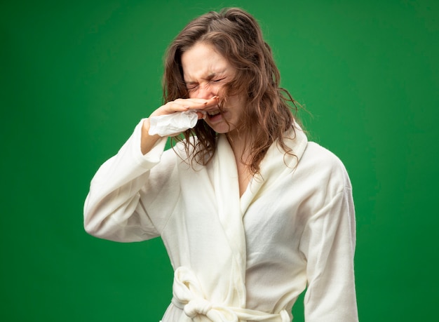 Foto gratuita niña enferma molesta con los ojos cerrados vistiendo una túnica blanca limpiando la nariz con una servilleta aislado en verde