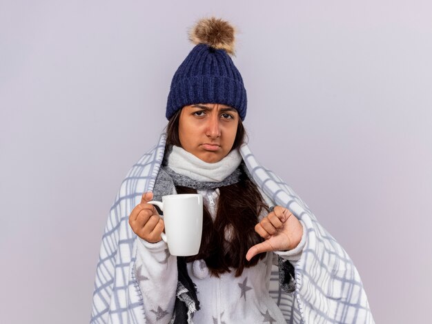 Niña enferma molesta con sombrero de invierno con bufanda envuelta en cuadros sosteniendo una taza de té mostrando el pulgar hacia abajo